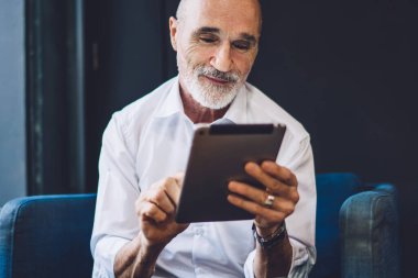 Gülümseyen, kel, resmi kıyafetler giyen, modern tableti zevkle karıştıran, Cafe 'deki koltukta oturan yaşlı adama sevindim.