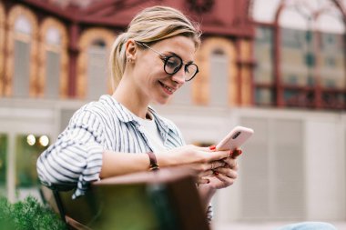 Sarışın, gülümseyen bir kadın, yeşil çalıların arkasında oturuyor, bacak bacak üstüne atıyor ve binanın bulanık arka planında akıllı telefon kullanıyor.