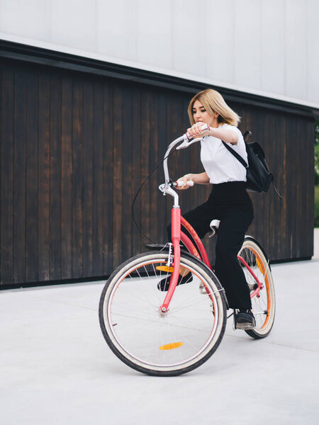 Adult blonde woman in causal outfit and with black backpack riding vintage pink bicycle against building in downtown looking away