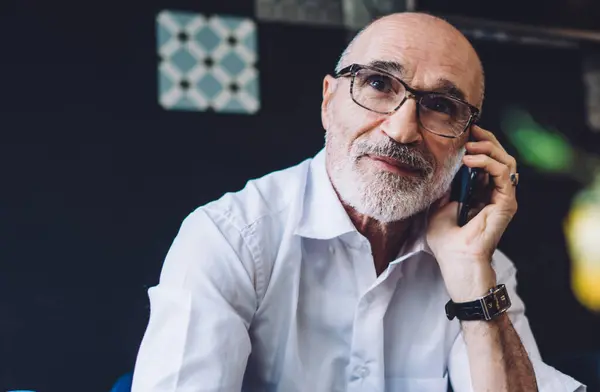Bald gray bearded male with eyeglasses in white shirt sitting at table on blue chair and looking away while speaking on phone