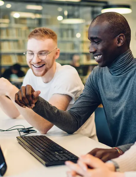 Happy multiethnic young people laughing and looking at screen while browsing computer with headphones and sitting at desk in modern library