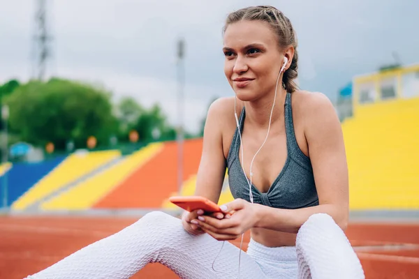 Adult athletic fit female in modern sportive wear using earbuds while browsing mobile sitting on ground of track of city stadium against grandstand having rest looking away