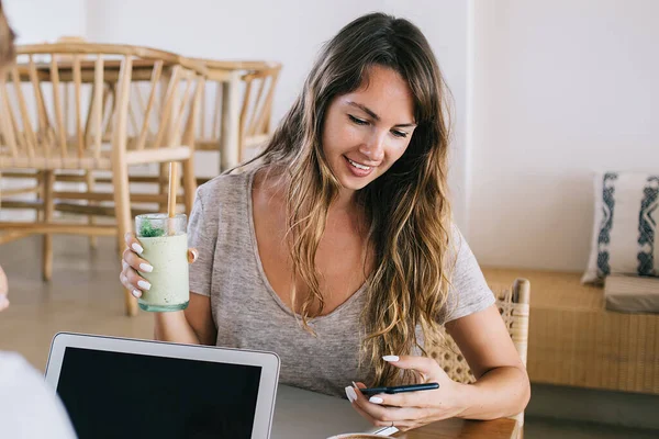 Young woman with cocktail and brown hair sitting at wooden desk and surfing on smartphone while smiling near man typing on laptop with black blank space in cafe