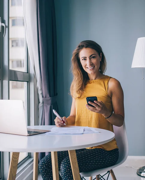 Glad young woman in yellow sleeveless blouse sitting at white round table with laptop and paper typing on smartphone at home and looking at camera