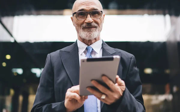 Handsome bearded old bald male in formal outfit and eyeglasses browsing tablet in front of business center on street looking at camera