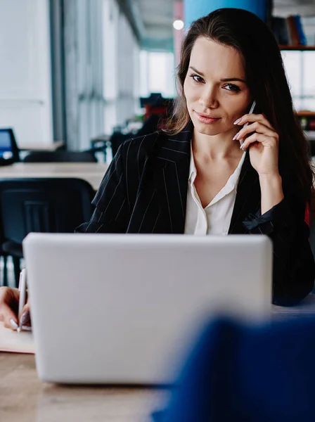 Pensive caucasian woman in formal wear having mobile phone talk writing information during working process, serious female employee sitting at desktop with notebook making cellphone call for banking