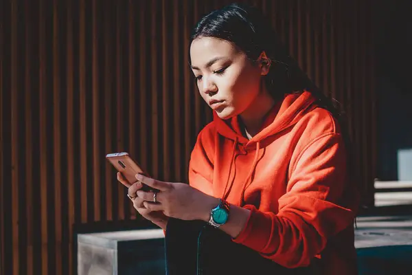 Chinese female blogger updating software on modern cellular phone while spending time on urban setting and connecting to public wifi internet, asian hipster girl checking notification for application