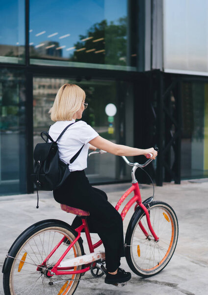 Back view of blonde female student dressed in casual clothes with black backpack sitting on bicycle with red frame while cycling on street