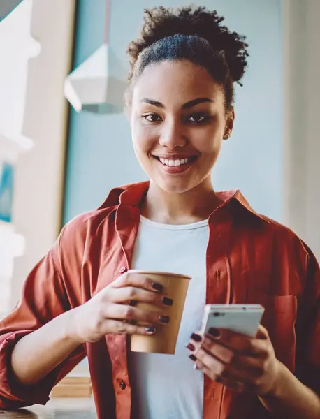 Half length portrait of cheerful female teenager with cellular phone in hand smiling at camera during coffee time in cafeteria, successful black woman in casual wear holding cup with caffeine beverage