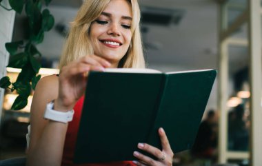 Siyah kitap okuyan ve kafede otururken ilginç komik hikayelerin tadını çıkaran neşeli, modern sarışın bir kadın.