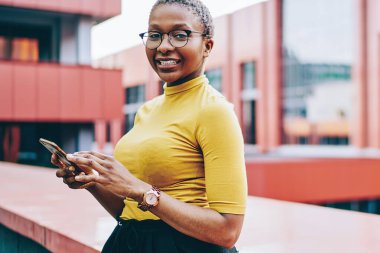 Neşeli Afro-Amerikan kadın portresi akıllı telefon kullanarak kentsel arka planda duruyor ve sohbet için cep telefonu tutan pozitif, koyu tenli kadın blogcu 4G bağlantısı var.