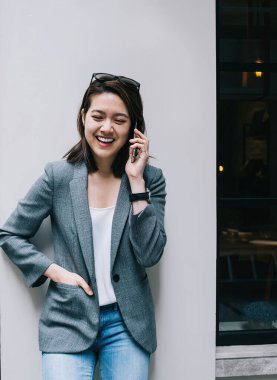 Güzel, neşeli, resmi giyinmiş genç bir kadın akıllı telefonuyla konuşuyor. Gözleri kapalı, neşeyle gülüyor şehir sokaklarında.