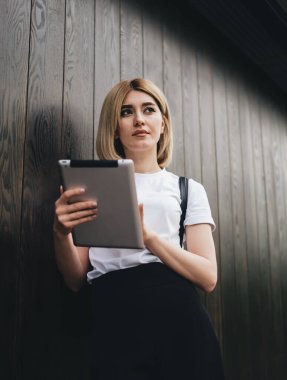 Ahşap duvara yaslanıp tablet kullanırken dalgın ve çekici bir kız öğrencinin dalgın bakışları.