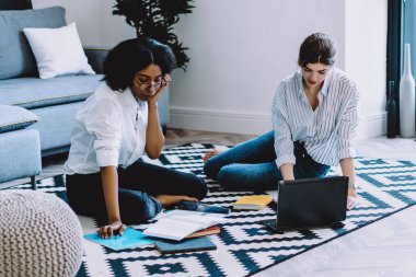 Kalifiye Afro-Amerikan ve Kafkasyalı öğrenciler yerde oturup laptop bilgisayar ve kablosuz internet kullanarak modern stil apartmanda ortak yayın üzerinde işbirliği yapıyorlar.