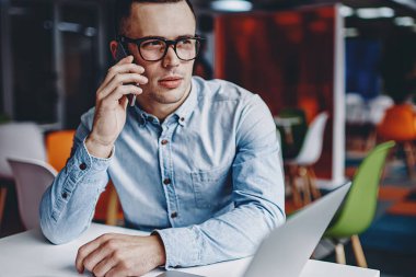 Gözlüklü dalgın genç adam akıllı telefondan arıyor ve ofiste bilgisayarla masada oturuyor. Netbook 'taki çalışma sürecinde cep telefonuyla iletişim kuran öğrenci.