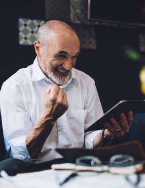 Beyaz tişörtlü, beyaz sakallı mutlu kel adam oturma odasında otururken yumruğunu kaldırmış tablet ekranına bakıyor.