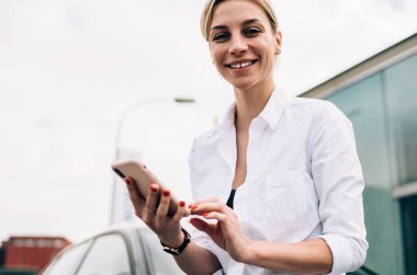 Güzel, neşeli sarışın kadın, günlük kıyafetlerle gülümseyen ve kameraya bakan, araca yaslanan ve otoparkta akıllı telefon kullanan.