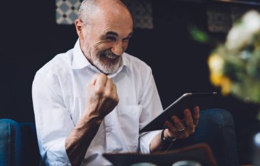 Beyaz tişörtlü, beyaz sakallı mutlu kel adam oturma odasında otururken yumruğunu kaldırmış tablet ekranına bakıyor.