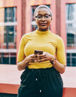 Sıradan giyinmiş Afro-Amerikan kadınlarının yarım boy portresi açık hava blogları için akıllı telefon tutuyor, pozitif esmer tenli hippi kız internetten cep telefonu sohbeti kullanarak kameraya bakıyor.