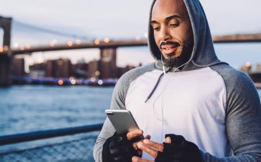 Genç Afro-Amerikalı adam ayakta durup kulaklıkla müzik dinlerken akıllı telefondan mesaj atıyor ve nehir ve köprünün arka planına hafifçe gülümsüyor.