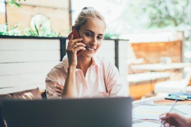 Akıllı telefondan konuşan ve bilgisayarın ekranına bakan hoş bir kadın. Meslektaşlar masada oturup dinlerken ve not defteri ve cihaz kullanarak not alırken.