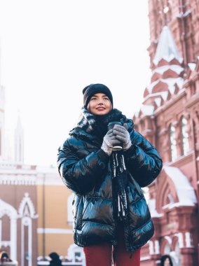 Koyu renk kışlık ceketli tatmin olmuş genç bir kadın açık gökyüzünde plastik bardakta sıcak kahveyle tarihi binaların yanında duruyor.