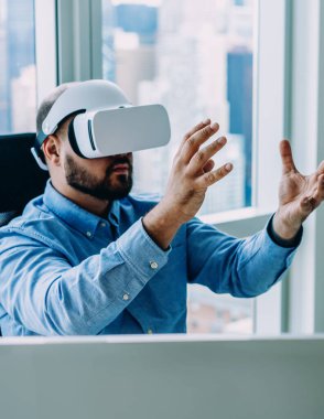 Yetişkin sakallı bir adam bilgisayarla masada otururken ve ofiste çalışırken modern sanal gerçeklik bardaklarında el kol hareketi yapıyor.