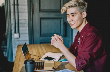 Sıradan giyinmiş pozitif Asyalı hippi öğrencilerin yaratıcı fikirleri üzerinde düşünmesi kendi geliştirme projesini geliştirmek için ahşap masaüstü bilgisayarında oturup üretken bir iş için dizüstü bilgisayarla düşünmesi.