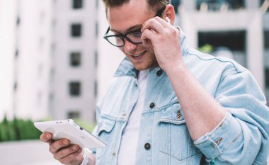 Gözlüklü ve kot ceketli yakışıklı adam tablet okuyor ve modern şehir sokaklarının bulanık arka planında mutlu bir şekilde gülümsüyor.