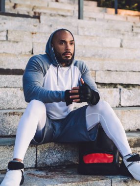 Genç, ciddi, Afro-Amerikalı spor elbiseli bir adam merdivenlerde oturuyor ve kulaklıkla müzik dinliyor.
