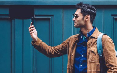 Sırt çantalı, modaya uygun giysileri ve güneş gözlükleriyle gülümseyen genç adam New York caddesinin mavi binasında dururken selfie çekiyor.