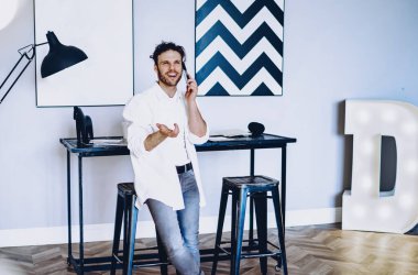 Günümüz modern tasarım odasında cep telefonuyla konuşan neşeli, beyaz tenli bir hippi. Cep telefonuyla konuşan mutlu ve yakışıklı bir adam.