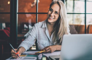 Gözlüklü, gülümseyen sarışın, kafede çalışırken masada dizüstü bilgisayar ve kağıt çizelgelerle oturuyor ve başka tarafa bakıyor.