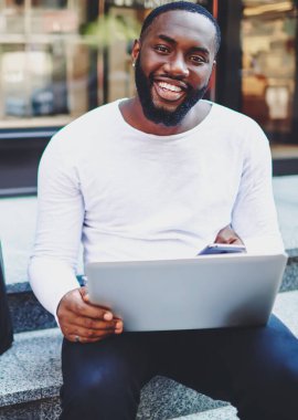 Modern akıllı telefon ve laptop cihazlarıyla merdivenlerde oturan mutlu zenci hipster portresi, neşeli Afro-Amerikan alışveriş koliği alışverişin ardından heyecanlanıyor, teknoloji kullanıcısı.