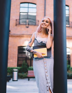 Not defterleri ile mutlu genç bir bayan. Gülümsüyor ve gözlerini kaçırıyor. Bir yandan da direğe yaslanıyor ve telefon çağrılarına cevap veriyor.