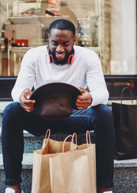 Alışveriş saatinden sonra merdivenlerde dinlenen, şık şapkalara bakan ve bir şeyler satın aldığı için kendini iyi hisseden, kağıt torbalı mutlu hippi erkek.