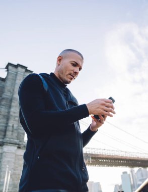 Aşağıdan keyifli, traşsız, siyah spor giysili genç adam şehir köprüsünde güpegündüz metal çitlerde akıllı telefonları dinliyor.