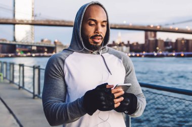 Genç kaslı Afrikalı Amerikalı adam ayakta durup kulaklıkla müzik dinlerken nehir ve köprünün arka planına akıllı telefondan mesaj atıyor.