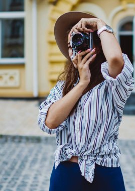 Arka planda fotoğraf çeken yetenekli kadın fotoğrafçı şehir ortamlarında dikiliyor, modaya uygun şapka ve giysili beyaz kadın şehirde fotoğraf çektirip eğlendiriyor.