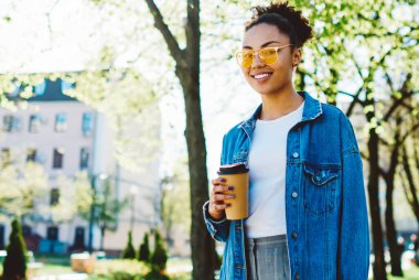Kot pantolon giymiş, kahve fincanı ile üniversiteye yürüyen ve kameraya gülümseyen modaya uygun güneş gözlüklü mutlu hippi kızın yarım boy Afro-Afrikalı kız portresi.