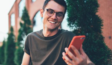 Görme düzeltmesi için gözlüklü neşeli erkek öğrenci akıllı telefon cihazıyla gülerek kampüste modern laptopla otururken, dışarıda serbest çalışan pozitif bir serbest oyuncu tarafından komik bir mesaj aldı.