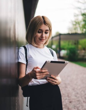 Beyaz tişörtlü modern sarışın kadın dışarıda dikilip ahşap bina duvarına yaslanırken tablet ve mesajlaşma yapıyor.