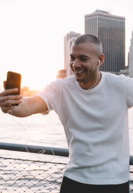 Kulaklıklı, akıllı telefonuyla gülümseyen ve selfie çeken genç yakışıklı etnik adam nehir ve şehrin zincire bağlı metal çitlere yaslanıyor.