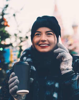 Rahat kışlık giysiler içinde mutlu bir genç kadın akıllı telefon kullanıyor ve bulanık arka planda, Noel sokağında sıcak kahve içiyor.