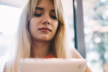 Düşük açılı, sarı saçlı, tablet kullanan, pencereye yaslanmış, rahat, modern bir kafede dinlenen bir kadın.