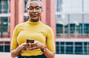 Sıradan giyinmiş Afro-Amerikan kadınlarının yarım boy portresi açık hava blogları için akıllı telefon tutuyor, pozitif esmer tenli hippi kız internetten cep telefonu sohbeti kullanarak kameraya bakıyor.