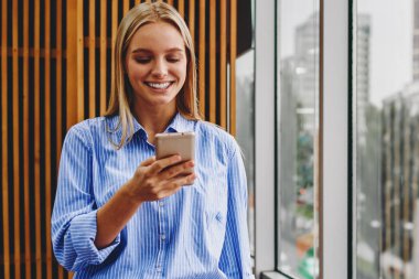 Neşeli kadın blogcu, ücretsiz 4G internetini kullanarak sosyal ağlarda modern akıllı telefon cihazında komik bildirimler okuyor. Ofis içinde cep telefonuyla sohbet eden olumlu genç kadın.