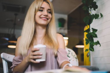 Alçak açılı, gülümseyen, açık pembe elbiseli, dizüstü bilgisayarda çalışan ve kahve dükkanının masasında bir fincan kahve tutan sarışın.