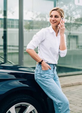 Şen şakrak sarışın bayan sürücü akıllı telefondan konuşuyor ve gülümsüyor. Siyah arabanın yanında duruyor ve şapkaya yaslanıyor.