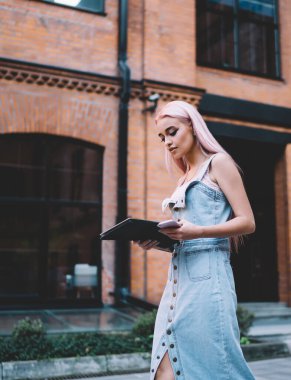 Uzun kotsuz, uzun kollu, ince sarışın kadın siyah klasör ve telefon taşıyan kırmızı tuğlalı bina boyunca sokakta yürüyor.